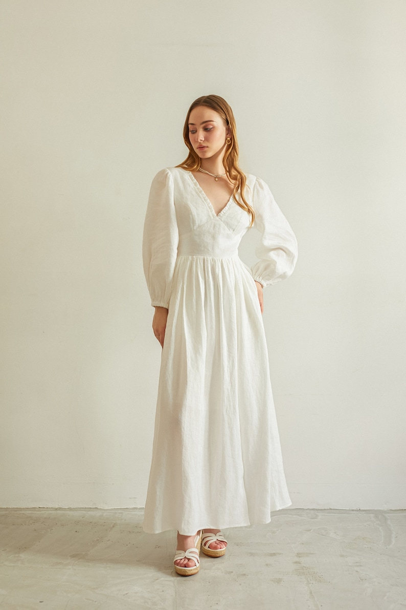 May include: A white linen maxi dress with a V-neckline, long sleeves, and a gathered skirt. The dress has a delicate lace trim around the neckline.