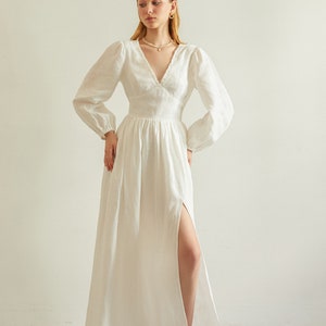 May include: A white linen maxi dress with a V-neckline, long sleeves, and a thigh-high slit. The dress has a fitted bodice and a flowing skirt.
