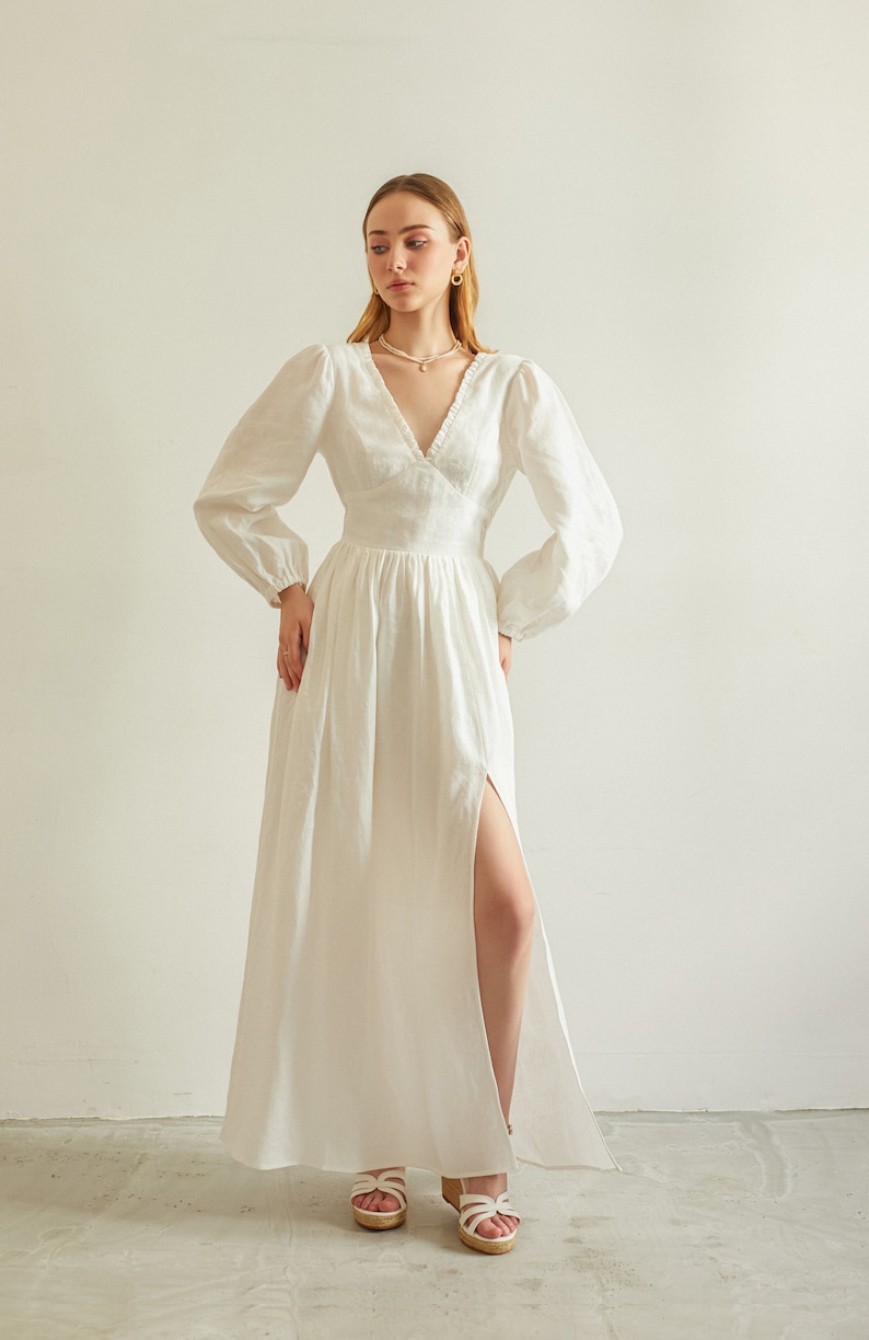 May include: A white linen maxi dress with a V-neckline, long sleeves, and a thigh-high slit. The dress has a fitted bodice and a flowing skirt.
