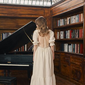 Puede incluir: Una mujer con un vestido blanco de espalda abierta y un dobladillo con volantes se encuentra en una habitaci&oacute;n con un piano de cola y estanter&iacute;as. El vestido tiene una espalda con cordones y mangas cortas y abullonadas.