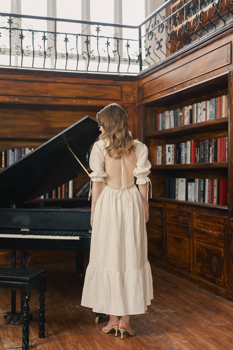 Puede incluir: Una mujer con un vestido blanco de espalda abierta y un dobladillo con volantes se encuentra en una habitaci&oacute;n con un piano de cola y estanter&iacute;as. El vestido tiene una espalda con cordones y mangas cortas y abullonadas.
