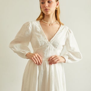 May include: A white linen dress with a v-neckline, long sleeves, and a ruffled trim. The dress has a fitted bodice and a flowing skirt with a side slit.