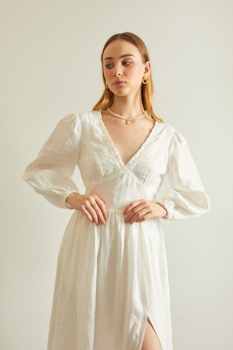 May include: A white linen dress with a v-neckline, long sleeves, and a ruffled trim. The dress has a fitted bodice and a flowing skirt with a side slit.