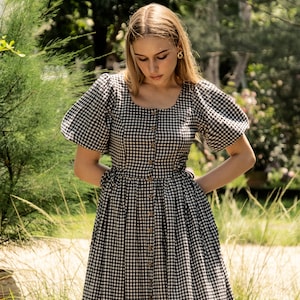 Puede incluir: Una mujer lleva un vestido de cuadros negros y blancos con mangas abullonadas y cierre de botones. El vestido tiene un corpi&ntilde;o ajustado y una falda fluida.