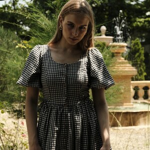 Puede incluir: Una mujer lleva un vestido de cuadros negros y blancos con mangas abullonadas. El vestido tiene un cierre de botones en la parte delantera y una falda fruncida.
