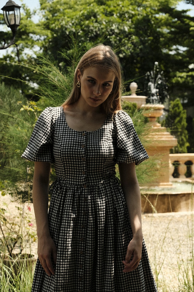 Puede incluir: Una mujer lleva un vestido de cuadros negros y blancos con mangas abullonadas. El vestido tiene un cierre de botones en la parte delantera y una falda fruncida.