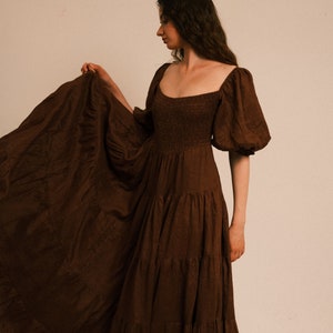 Pode incluir: Uma mulher vestindo um vestido maxi marrom em camadas com mangas bufantes. O vestido tem um decote quadrado e um corpete justo.