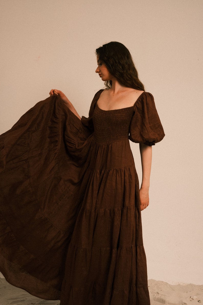 Pode incluir: Uma mulher vestindo um vestido maxi marrom em camadas com mangas bufantes. O vestido tem um decote quadrado e um corpete justo.