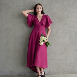 Puede incluir: Vestido magenta con escote en V y botones. Mangas cortas y acampanadas y dobladillo asimétrico. La persona sostiene rosas blancas y lleva zapatos negros. El vestido es de tela fluida.
