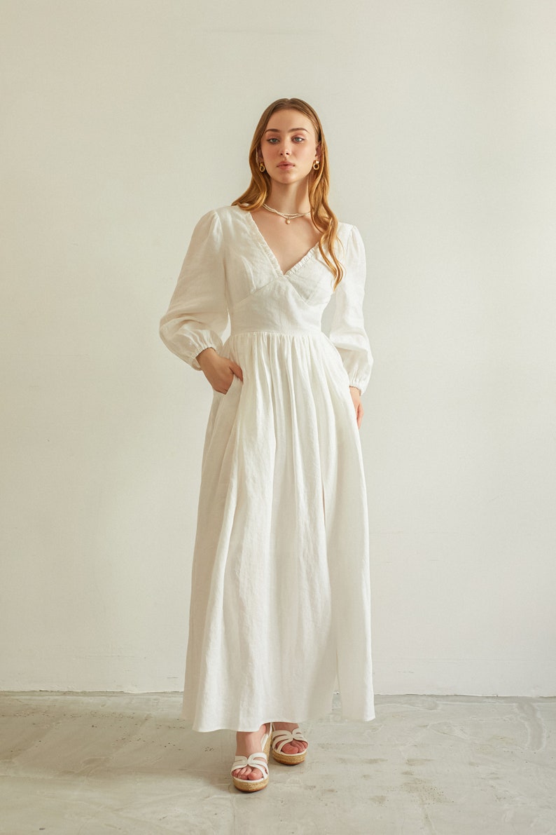 May include: A white linen maxi dress with a V-neckline, long sleeves, and a gathered skirt. The dress has a relaxed fit and a flowy silhouette.