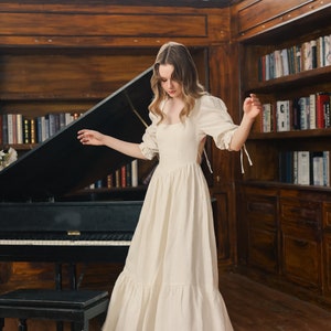 Puede incluir: Una mujer lleva un vestido blanco de lino con capas, escote cuadrado y espalda baja. El vestido tiene mangas abullonadas con lazos en las mu&ntilde;ecas. Est&aacute; de pie frente a un piano de cola negro con un taburete de madera.