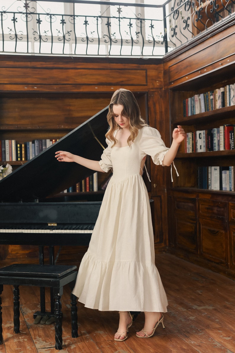 Puede incluir: Una mujer lleva un vestido blanco de lino con capas, escote cuadrado y espalda baja. El vestido tiene mangas abullonadas con lazos en las mu&ntilde;ecas. Est&aacute; de pie frente a un piano de cola negro con un taburete de madera.