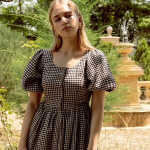 Puede incluir: Una mujer con un vestido de cuadros negros y blancos con mangas abullonadas y cierre de botones. Est&aacute; de pie en un jard&iacute;n con una fuente en el fondo.