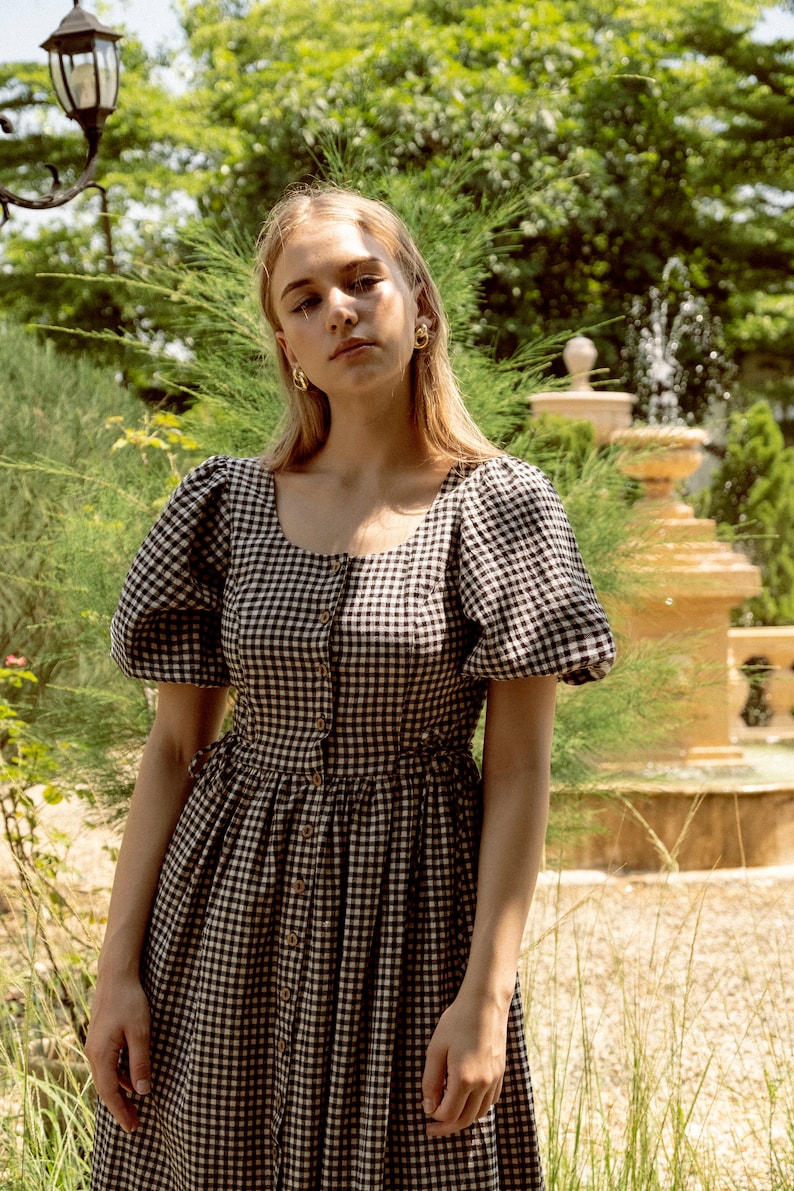 Puede incluir: Una mujer con un vestido de cuadros negros y blancos con mangas abullonadas y cierre de botones. Est&aacute; de pie en un jard&iacute;n con una fuente en el fondo.