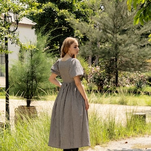 Puede incluir: Una mujer con un vestido de cuadros negros y blancos con mangas abullonadas y botas negras. Est&aacute; de pie en un jard&iacute;n con hierba verde y &aacute;rboles en el fondo.