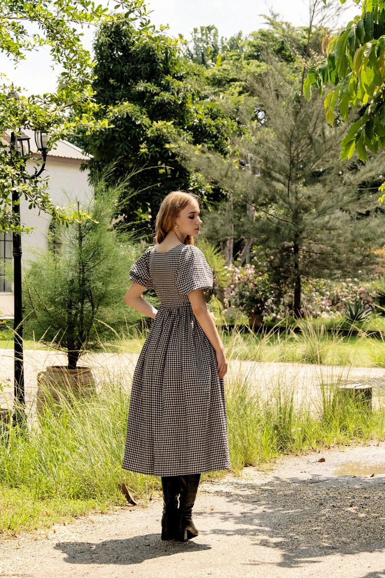 Puede incluir: Una mujer con un vestido de cuadros negros y blancos con mangas abullonadas y botas negras. Est&aacute; de pie en un jard&iacute;n con hierba verde y &aacute;rboles en el fondo.