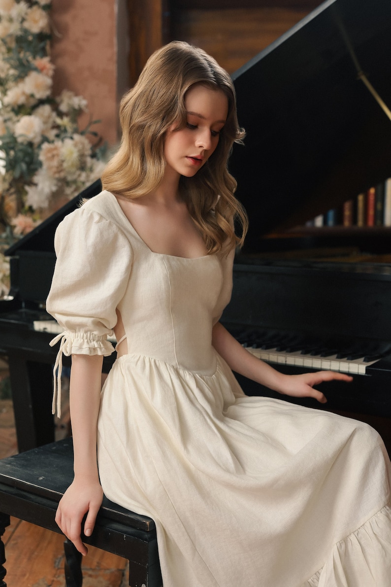 Puede incluir: Una mujer con un vestido blanco de lino con mangas abullonadas se sienta en un piano de cola negro. El vestido tiene un escote cuadrado y un corpi&ntilde;o ajustado. La mujer tiene el pelo largo y rubio y est&aacute; mirando las teclas del piano.