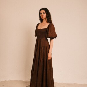 Pode incluir: Uma mulher vestindo um vestido maxi marrom em camadas com mangas bufantes e um decote quadrado. O vestido tem um corpete franzido e &eacute; feito de um tecido leve. Ela est&aacute; em p&eacute; em uma superf&iacute;cie arenosa.