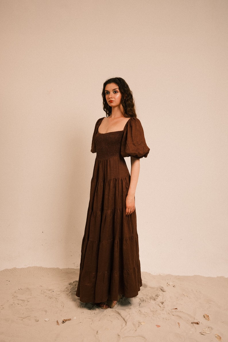 Pode incluir: Uma mulher vestindo um vestido maxi marrom em camadas com mangas bufantes e um decote quadrado. O vestido tem um corpete franzido e &eacute; feito de um tecido leve. Ela est&aacute; em p&eacute; em uma superf&iacute;cie arenosa.