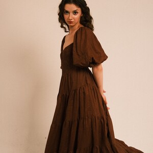 Pode incluir: Uma mulher vestindo um vestido maxi marrom em camadas com mangas bufantes. O vestido tem um decote quadrado e &eacute; feito de um tecido macio e fluido.