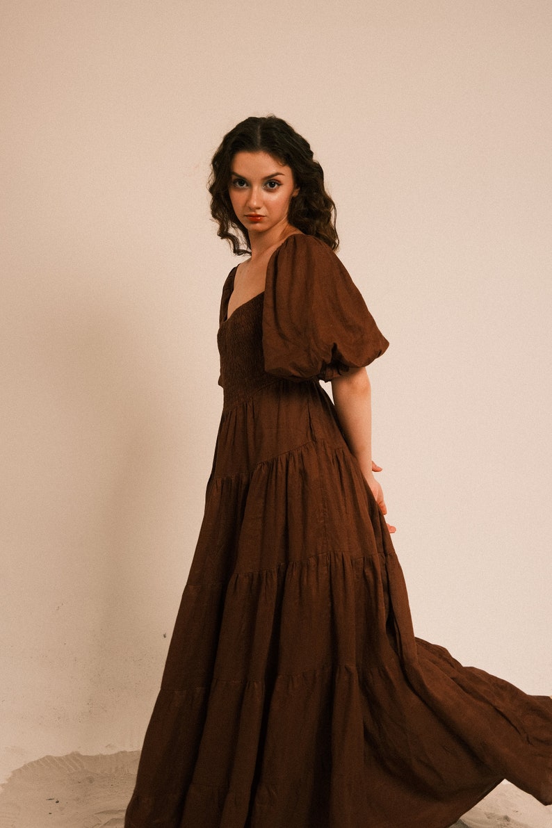 Pode incluir: Uma mulher vestindo um vestido maxi marrom em camadas com mangas bufantes. O vestido tem um decote quadrado e &eacute; feito de um tecido macio e fluido.