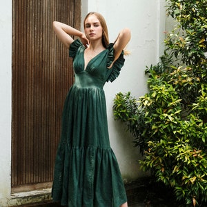 May include: A woman wearing a green tiered maxi dress with ruffled sleeves and white sandals. She is standing in front of a wooden door and a white wall.
