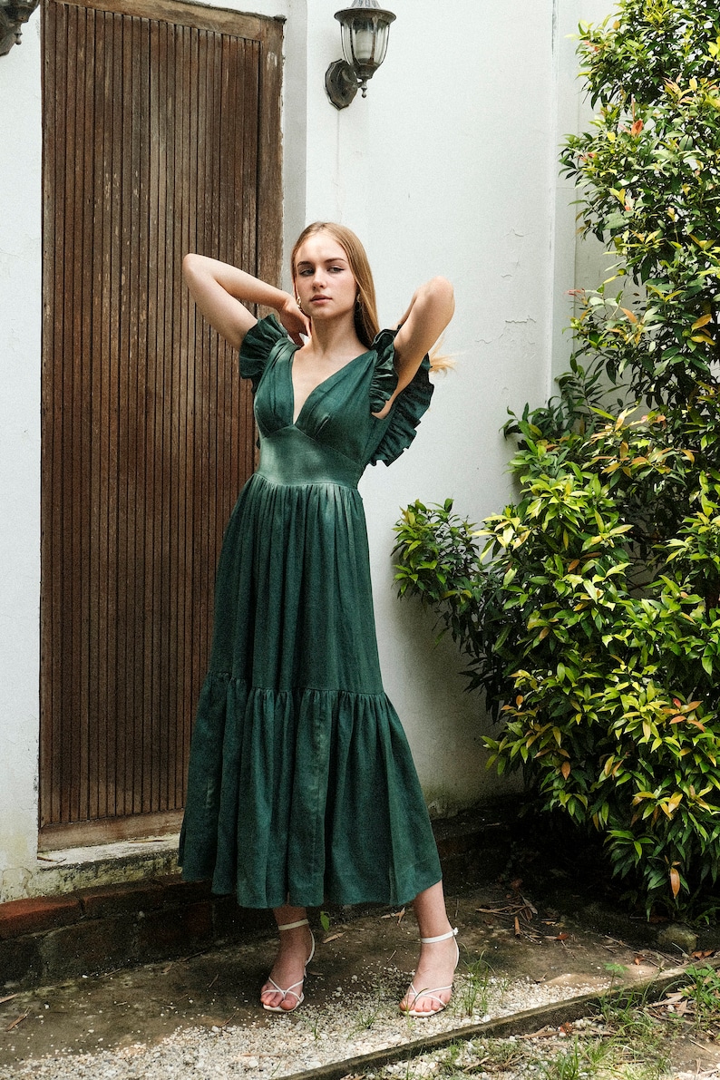 May include: A woman wearing a green tiered maxi dress with ruffled sleeves and white sandals. She is standing in front of a wooden door and a white wall.