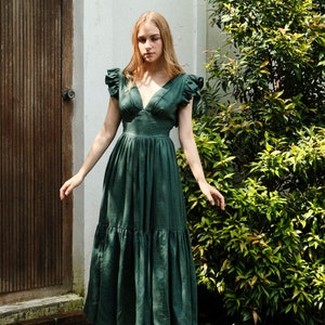 May include: A woman wearing a green linen dress with ruffled sleeves and a v-neckline. The dress has a tiered skirt and is midi length. She is wearing white sandals.