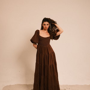 Pode incluir: Uma mulher vestindo um vestido maxi marrom em camadas com mangas bufantes, em p&eacute; em uma superf&iacute;cie arenosa.