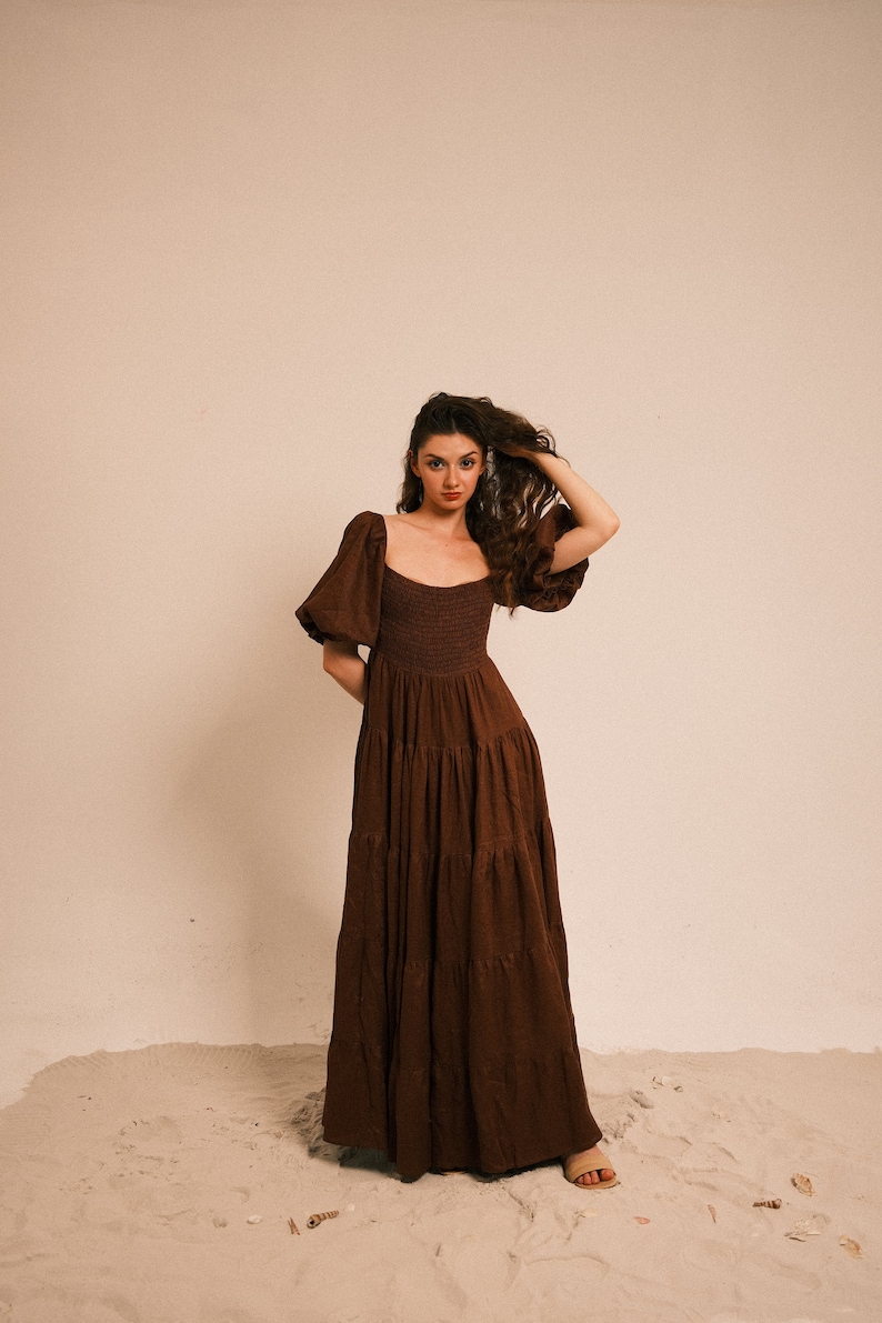 Pode incluir: Uma mulher vestindo um vestido maxi marrom em camadas com mangas bufantes, em p&eacute; em uma superf&iacute;cie arenosa.