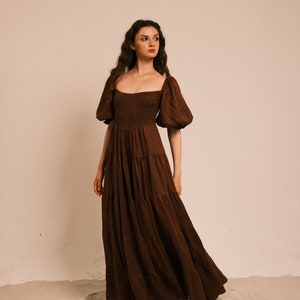 Pode incluir: Uma mulher vestindo um vestido maxi marrom em camadas com mangas bufantes e decote quadrado. O vestido &eacute; feito de um tecido macio e fluido e tem um caimento solto. A mulher est&aacute; em p&eacute; em uma superf&iacute;cie arenosa.