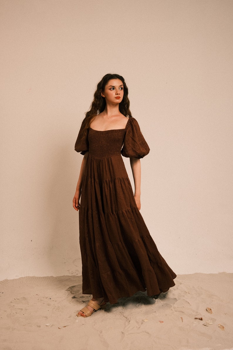 Pode incluir: Uma mulher vestindo um vestido maxi marrom em camadas com mangas bufantes e decote quadrado. O vestido &eacute; feito de um tecido macio e fluido e tem um caimento solto. A mulher est&aacute; em p&eacute; em uma superf&iacute;cie arenosa.