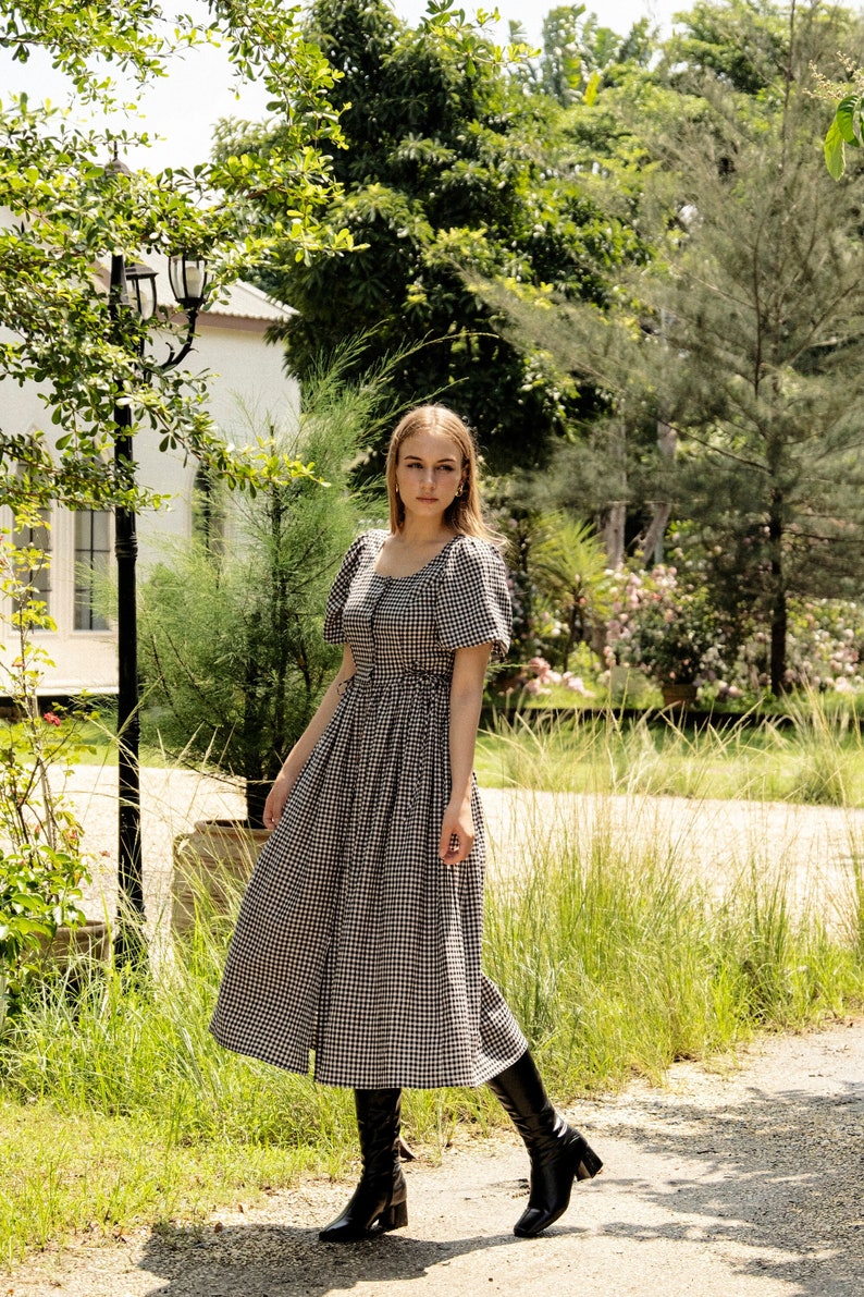 Puede incluir: Una mujer con un vestido midi de cuadros vichy blanco y negro con mangas abullonadas y botas negras de ca&ntilde;a alta. Est&aacute; de pie en un camino en un jard&iacute;n.