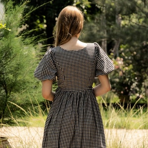 Puede incluir: Un vestido de cuadros vichy blanco y negro con escote cuadrado y mangas abullonadas. El vestido es midi y tiene una falda fruncida.