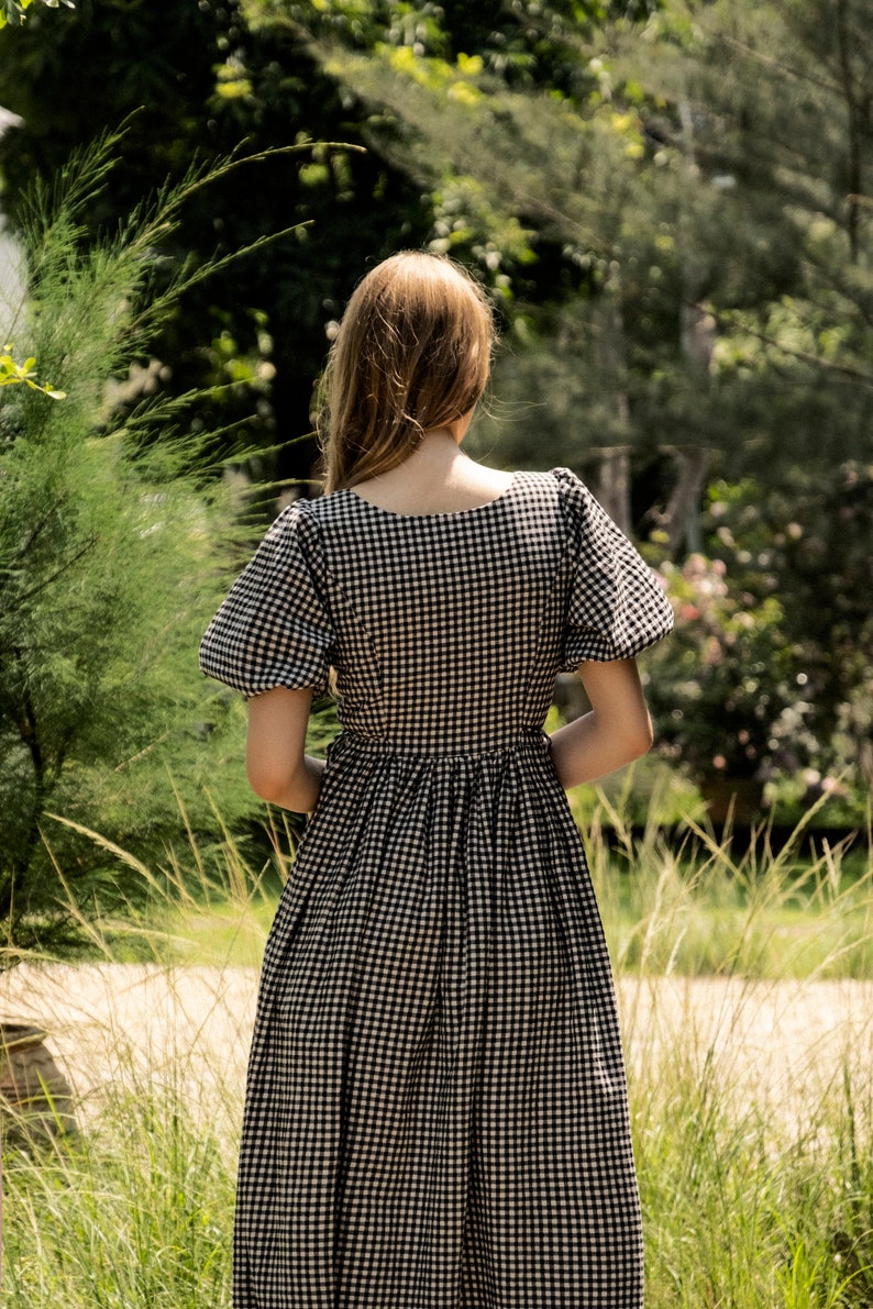Puede incluir: Un vestido de cuadros vichy blanco y negro con escote cuadrado y mangas abullonadas. El vestido es midi y tiene una falda fruncida.