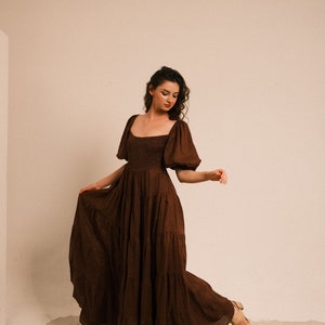 Pode incluir: Uma mulher vestindo um vestido maxi marrom em camadas com mangas bufantes est&aacute; em p&eacute; em uma superf&iacute;cie arenosa. O vestido tem um decote quadrado e um corpete justo.