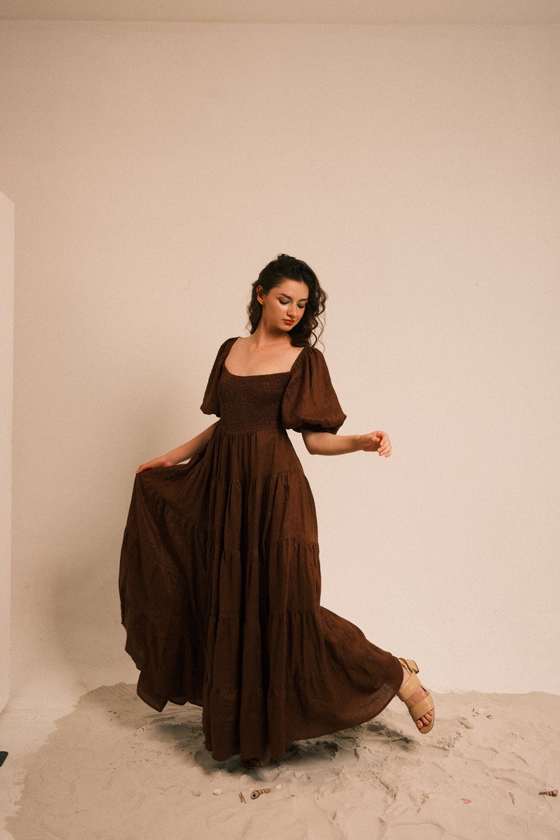Pode incluir: Uma mulher vestindo um vestido maxi marrom em camadas com mangas bufantes est&aacute; em p&eacute; em uma superf&iacute;cie arenosa. O vestido tem um decote quadrado e um corpete justo.