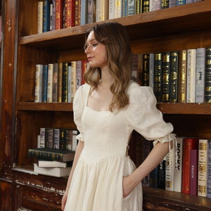 Puede incluir: Una mujer lleva un vestido blanco de lino con escote cuadrado y mangas abullonadas. El vestido tiene un detalle de corte en la espalda y un bolsillo en el lateral. Est&aacute; de pie frente a una estanter&iacute;a llena de libros.