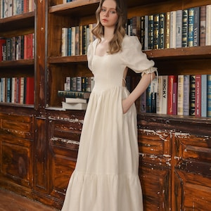Puede incluir: Un vestido blanco de lino con escote cuadrado, mangas abullonadas y falda de vuelo. El vestido tiene la espalda descubierta y un detalle de lazo en la cintura. La modelo est&aacute; de pie frente a una estanter&iacute;a.