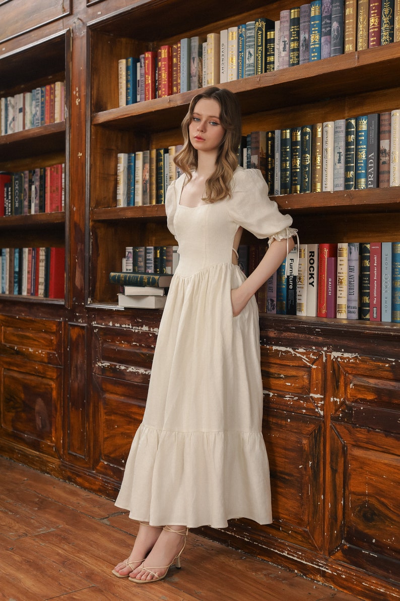 Puede incluir: Un vestido blanco de lino con escote cuadrado, mangas abullonadas y falda de vuelo. El vestido tiene la espalda descubierta y un detalle de lazo en la cintura. La modelo est&aacute; de pie frente a una estanter&iacute;a.