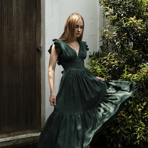 May include: A woman wearing a dark green, ruffled, sleeveless maxi dress with a deep V-neckline. She is standing in front of a white wall and a wooden door.