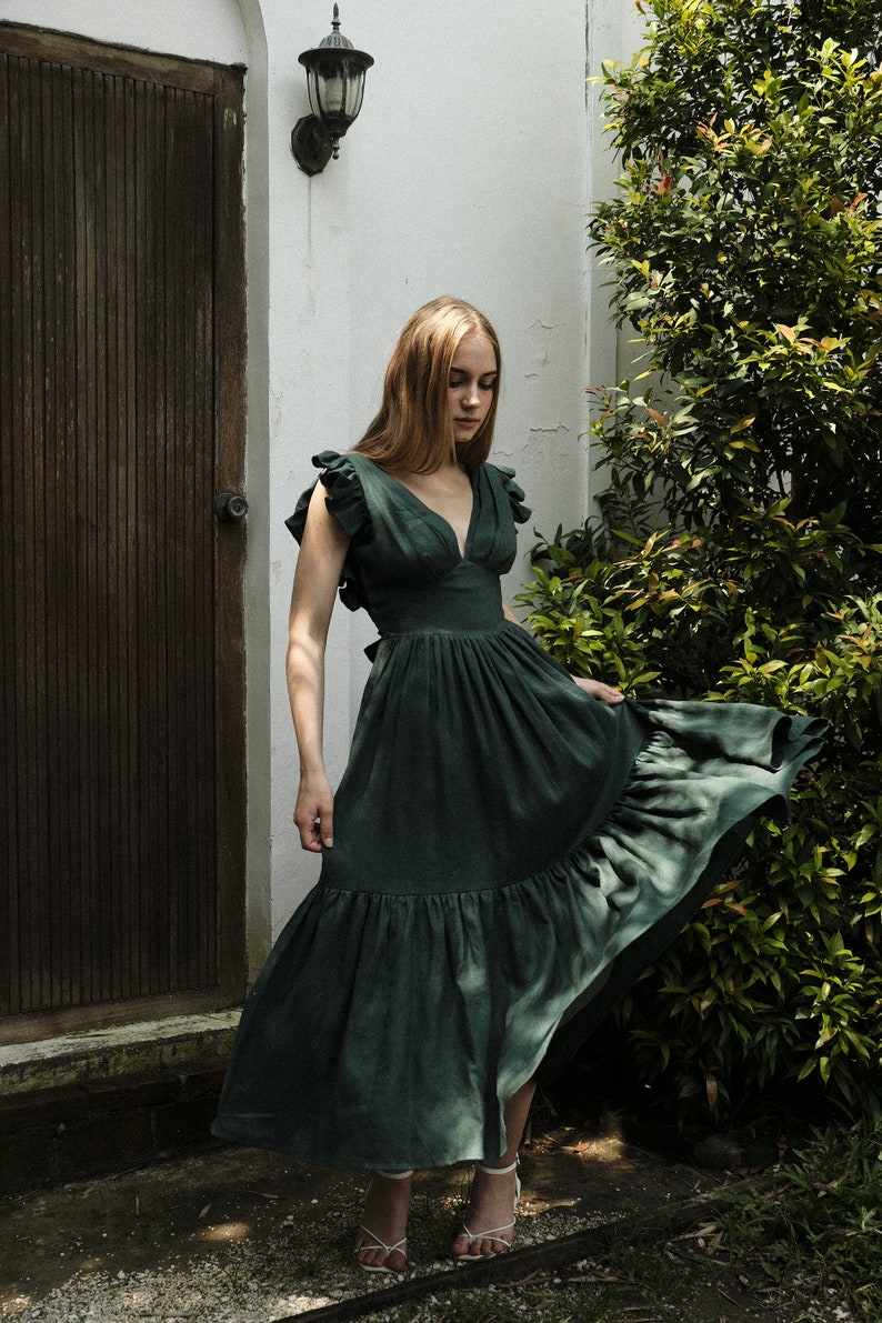 May include: A woman wearing a dark green, ruffled, sleeveless maxi dress with a deep V-neckline. She is standing in front of a white wall and a wooden door.