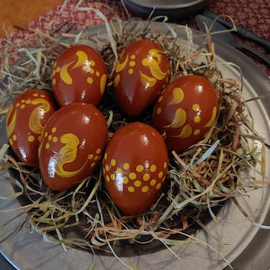 May include: Six brown Easter eggs with yellow painted designs are nestled in a bed of straw on a silver plate.