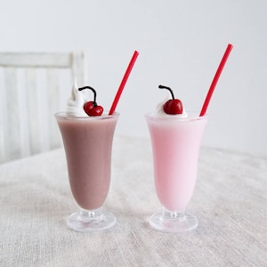 May include: Two miniature milkshakes with red straws and cherries on top. One milkshake is pink and the other is brown.