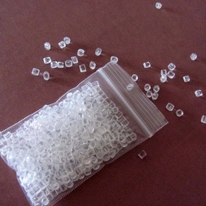 May include: A clear plastic bag filled with small, clear, square-shaped beads. The bag is open and some beads have spilled out onto a brown surface.