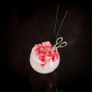 May include: A clear glass filled with crushed ice and a red liquid, topped with a pair of silver scissors and two black straws.