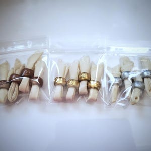 May include: Three clear plastic bags each containing multiple white dog chew bones with gold, bronze, and silver rings around them.