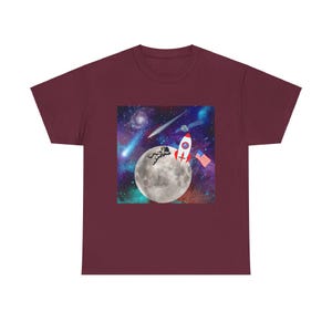 May include: Burgundy t-shirt featuring a colorful space-themed graphic. The design includes a full moon, a rocket, an astronaut, an American flag, and a starry galaxy background. The shirt is made of a soft, comfortable material.