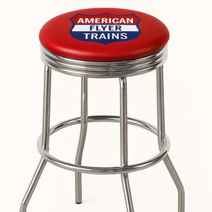 May include: A retro-style bar stool with a red seat featuring the "AMERICAN FLYER TRAINS" logo. The stool has a chrome-plated steel frame and a circular footrest. The seat is approximately 14 inches in diameter.