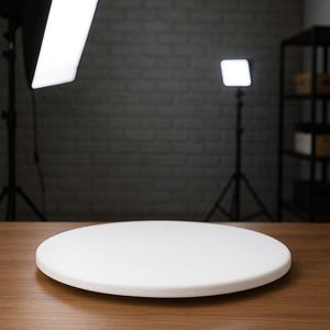 May include: A white, round ceramic pizza stone sits on a wooden surface. The stone is the focal point, with studio lighting and a brick wall in the background. The stone is a solid white color.